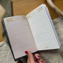 Load image into Gallery viewer, Open planner with a decorative cover held by a hand on a wooden surface