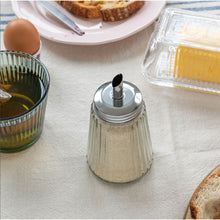 Load image into Gallery viewer, Silver salt shaker on a table with a glass of oil and bread slices in the background.