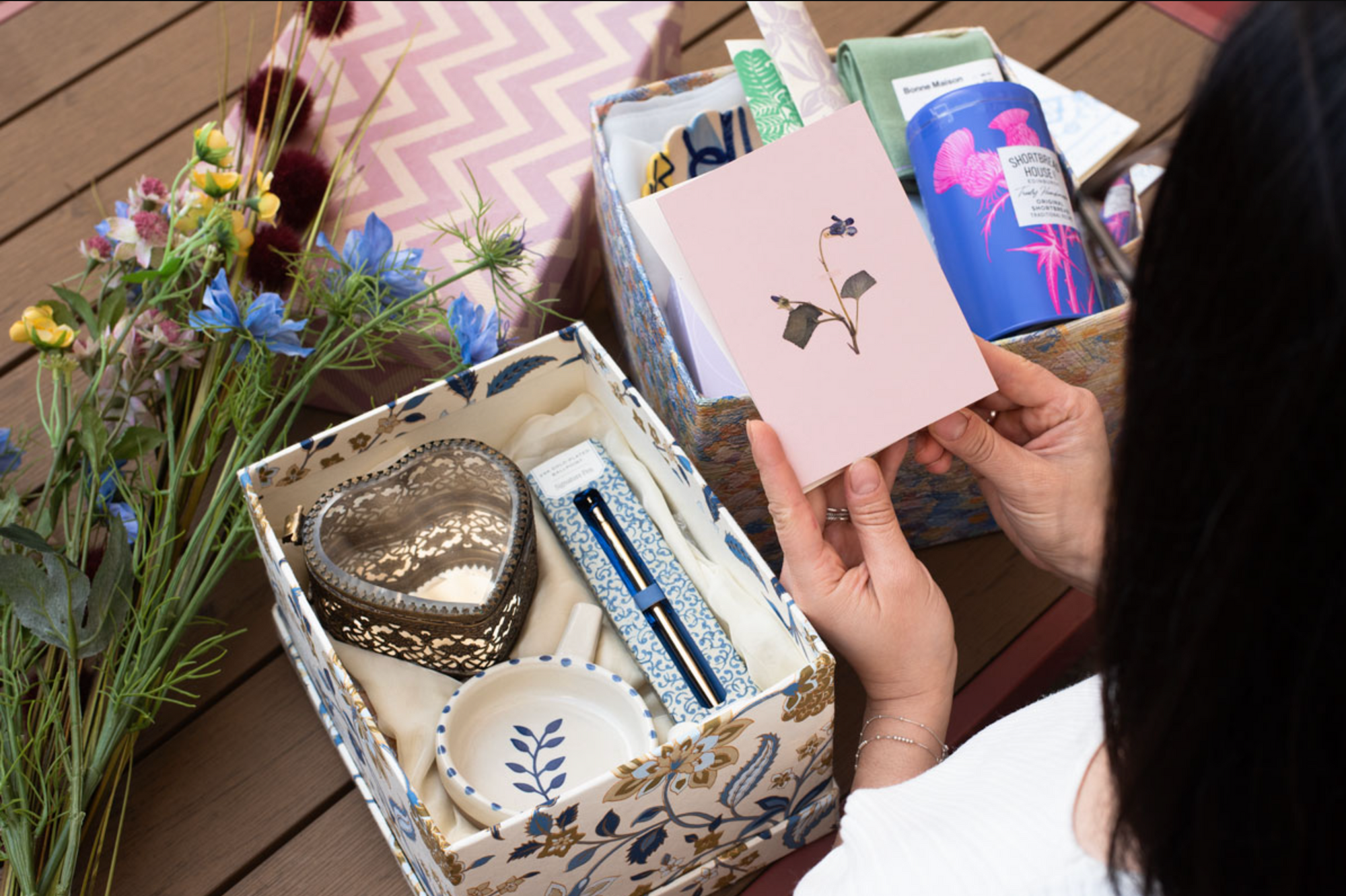 Person holding a card with a dry flower over a decorative box with stationery with flower motif and heart shapes items.