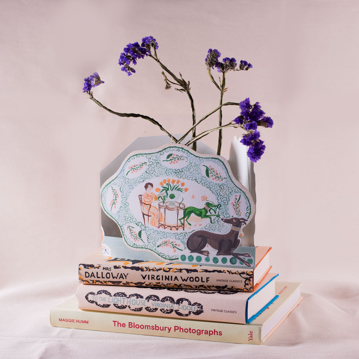 Decorative plate with a design of two people sitting together, a dog, and a horse, placed on stacked books with purple flowers in the background.