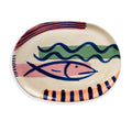 An oval ceramic serving platter with a hand-painted Mediterranean style design featuring a pink fish, blue waves, and striped accents on a cream background.